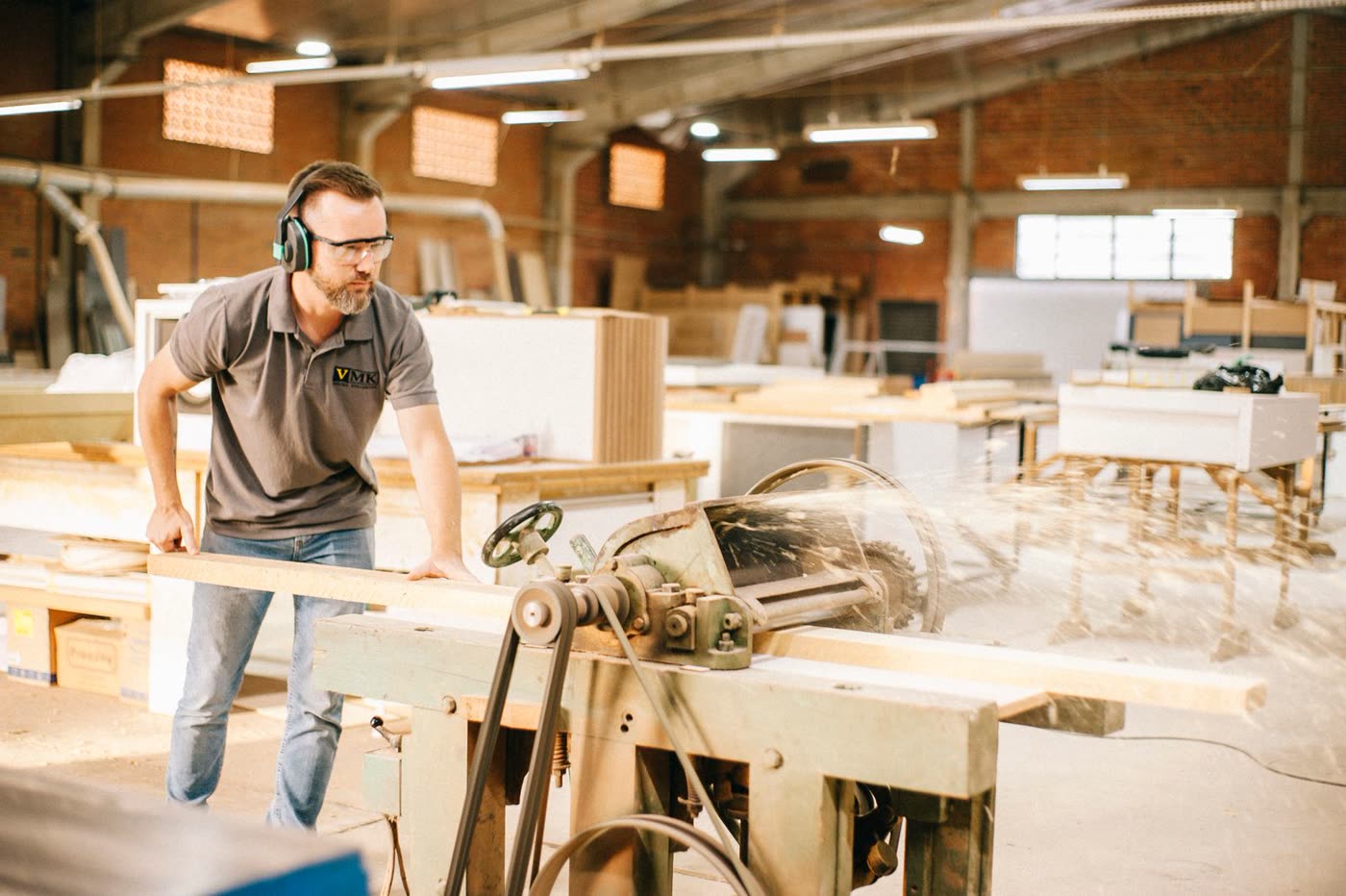 A craftsman in safety glasses and ear protection feeds a wood plank through an industrial planer in a bright woodshop