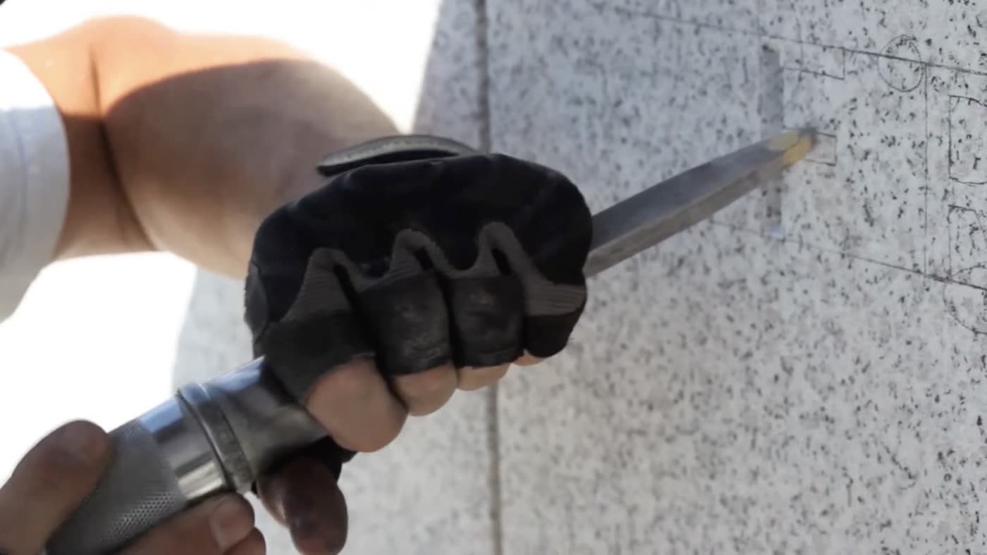 A gloved hand uses a chisel to engrave lettering into a granite slab