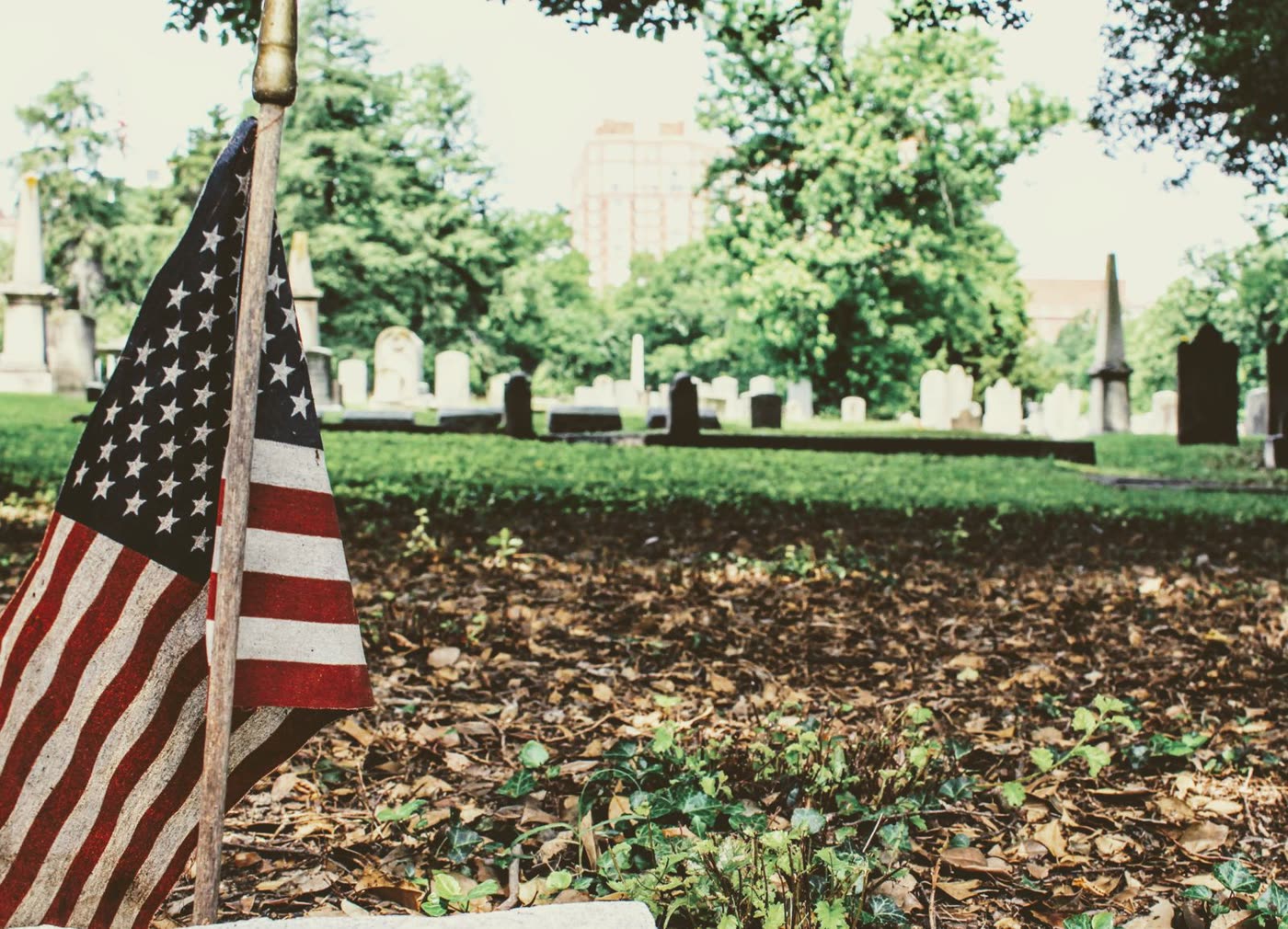 A small American flag on a stand beside headstones in a tree-shaded cemetery